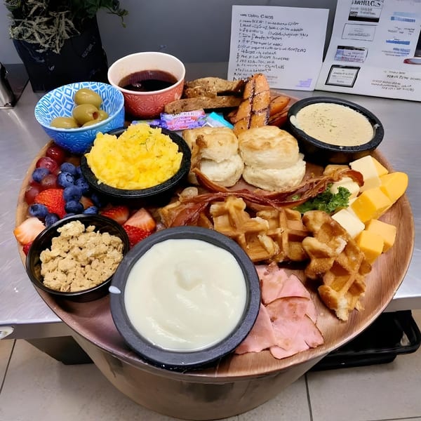 A large wooden platter with breakfast foods including waffles, bacon, fruit, cheese, and biscuits.
