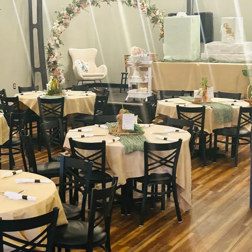 A decorated dining room with round tables, black chairs, and a floral arch for an event.