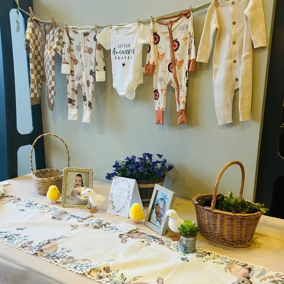 Baby clothes hang above a table decorated with baskets, framed photos, and chick figurines.