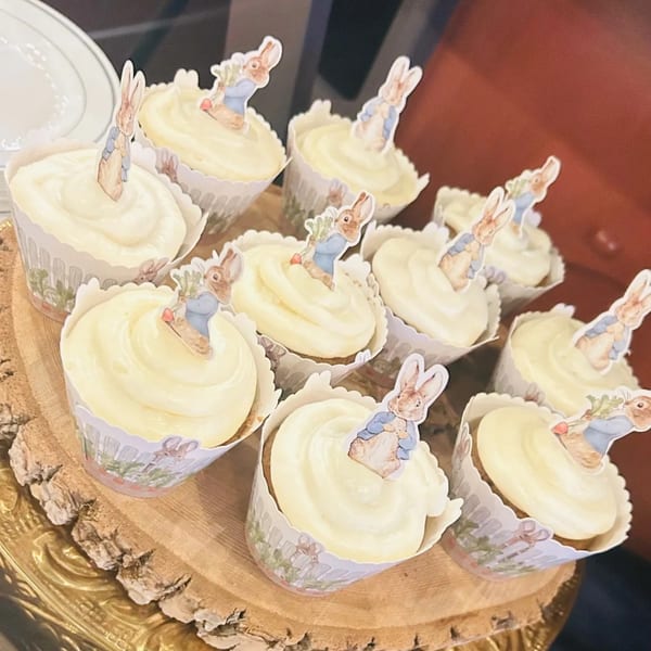 A wooden tray holds cupcakes with white frosting and Peter Rabbit toppers, ready for a party.