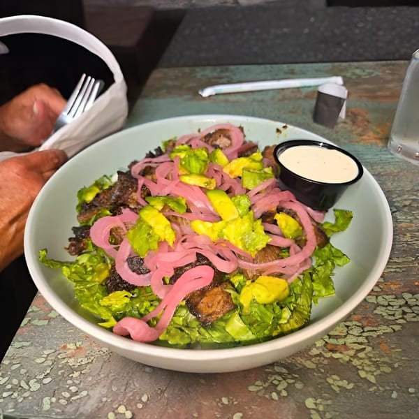 A vibrant steak salad with pickled red onions, avocado, and a side of dressing in a white bowl.