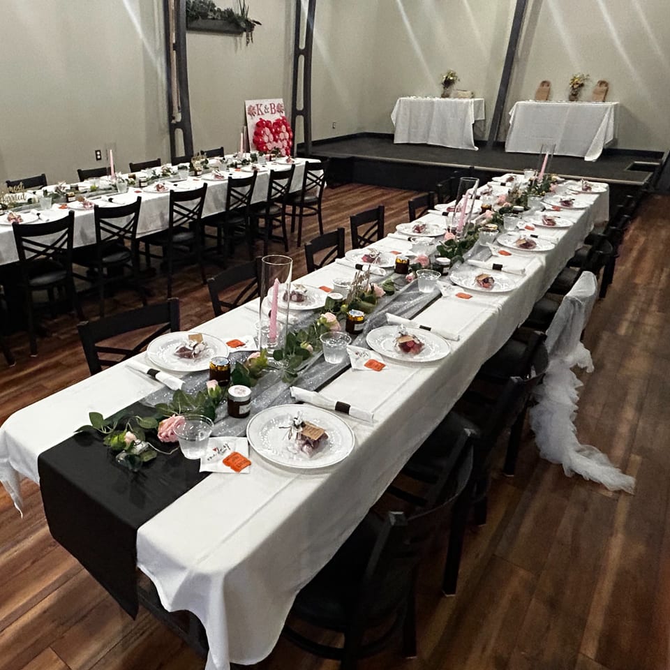 Elegant banquet hall with white-clothed tables, plated meals, floral centerpieces, black chairs, and a decorated stage.