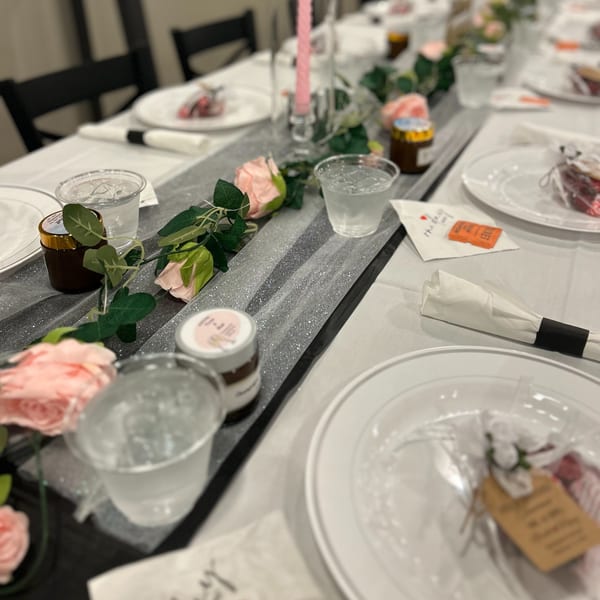Elegant dining table set with white plates, glasses, napkins, floral greenery, and small jars arranged along a glittering runner.